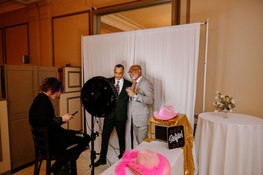 Two men pose for a photo booth photographer at an elegant event venue with white drape backdrop.