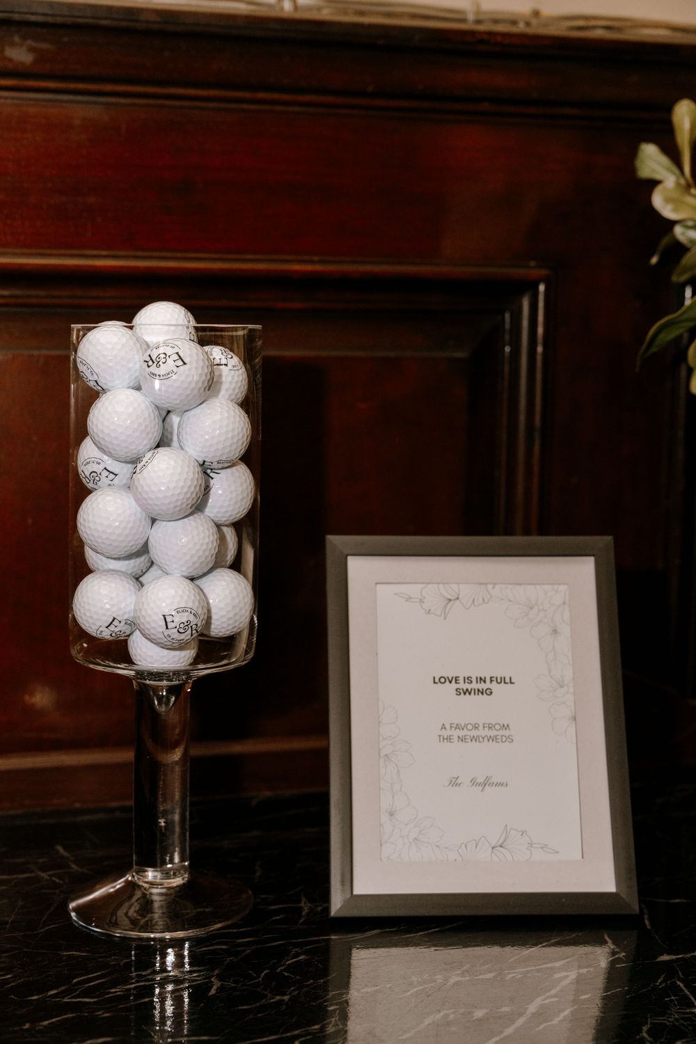 Glass vase filled with monogrammed golf balls beside a framed wedding favor sign on black marble.