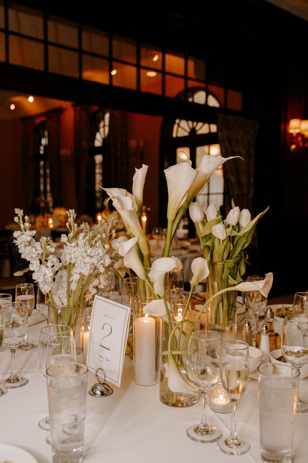 Elegant wedding reception table with white calla lilies, tulips, candles, and glassware on a white tablecloth.