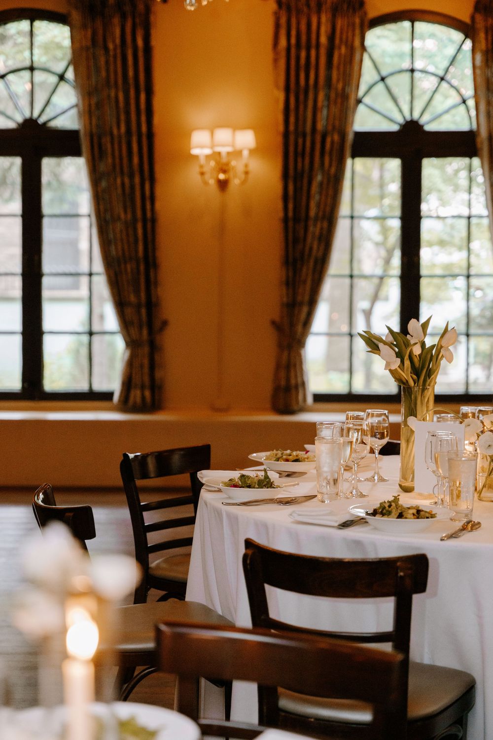 Romantic wedding reception dining room with candlelit tables, white linens, and arched windows.
