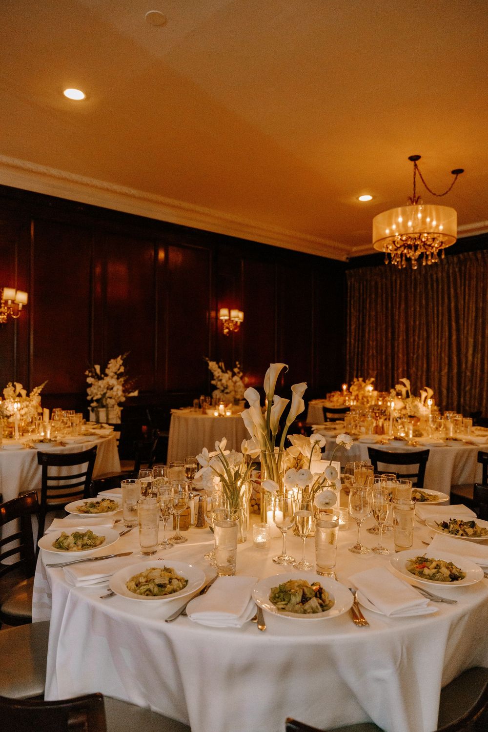 Elegant wedding reception room with white tablecloths, calla lily centerpieces, candlelight, and a warm chandelier glow.