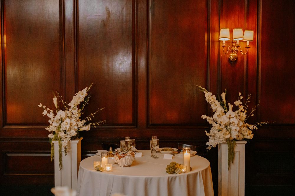 Elegant sweetheart table with white floral arrangements on pedestals and candlelight in a wood-paneled reception venue.