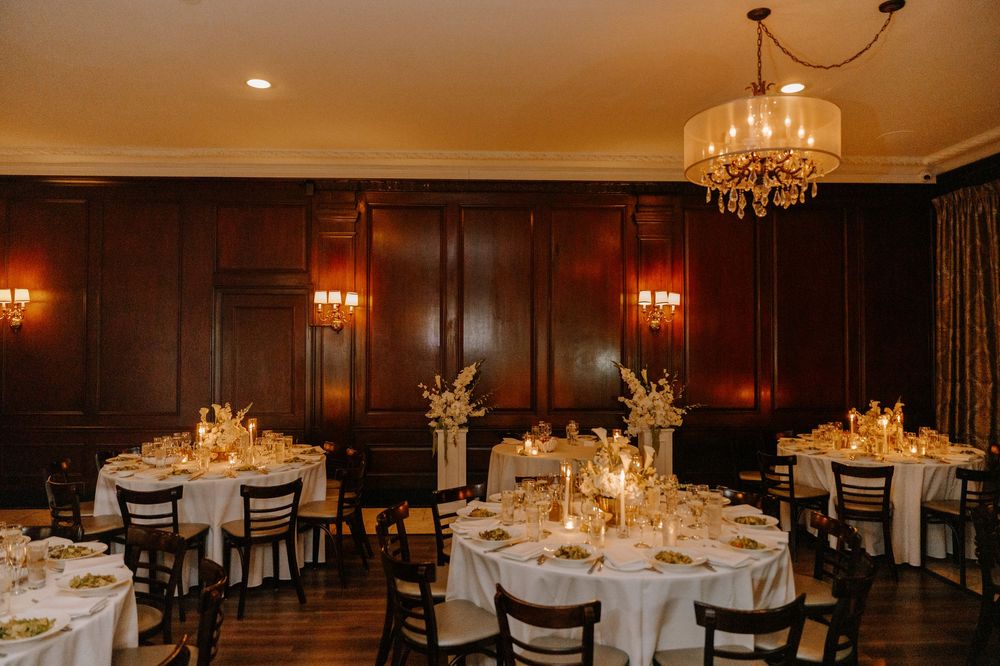 Elegant wedding reception room with round tables, white linens, candlelight, floral centerpieces, and warm wood-paneled walls.