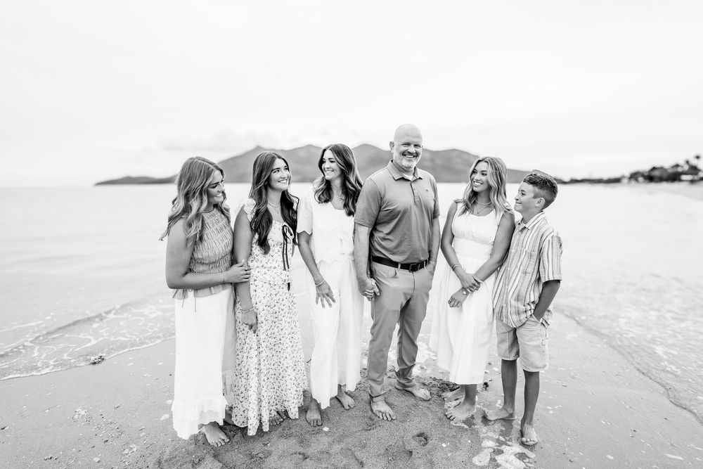 A joyful family of six poses together on a sandy beach with ocean and mountains in the background.