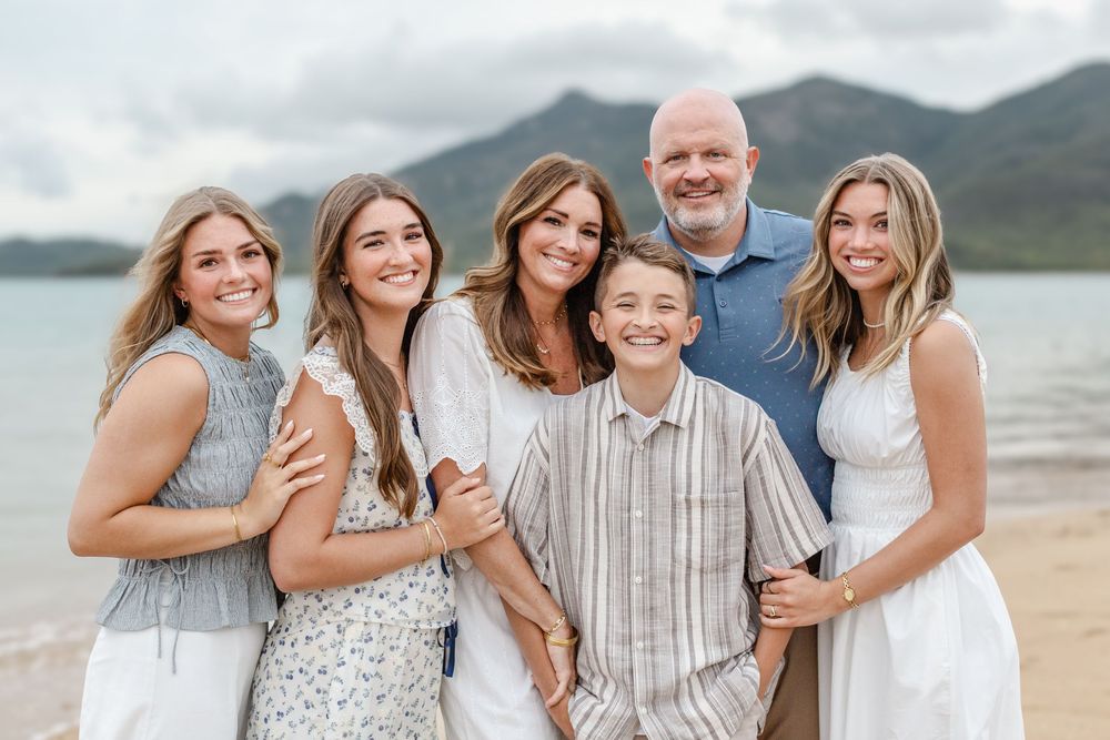 Family of six smiling together on a beach with mountain backdrop on a cloudy day.