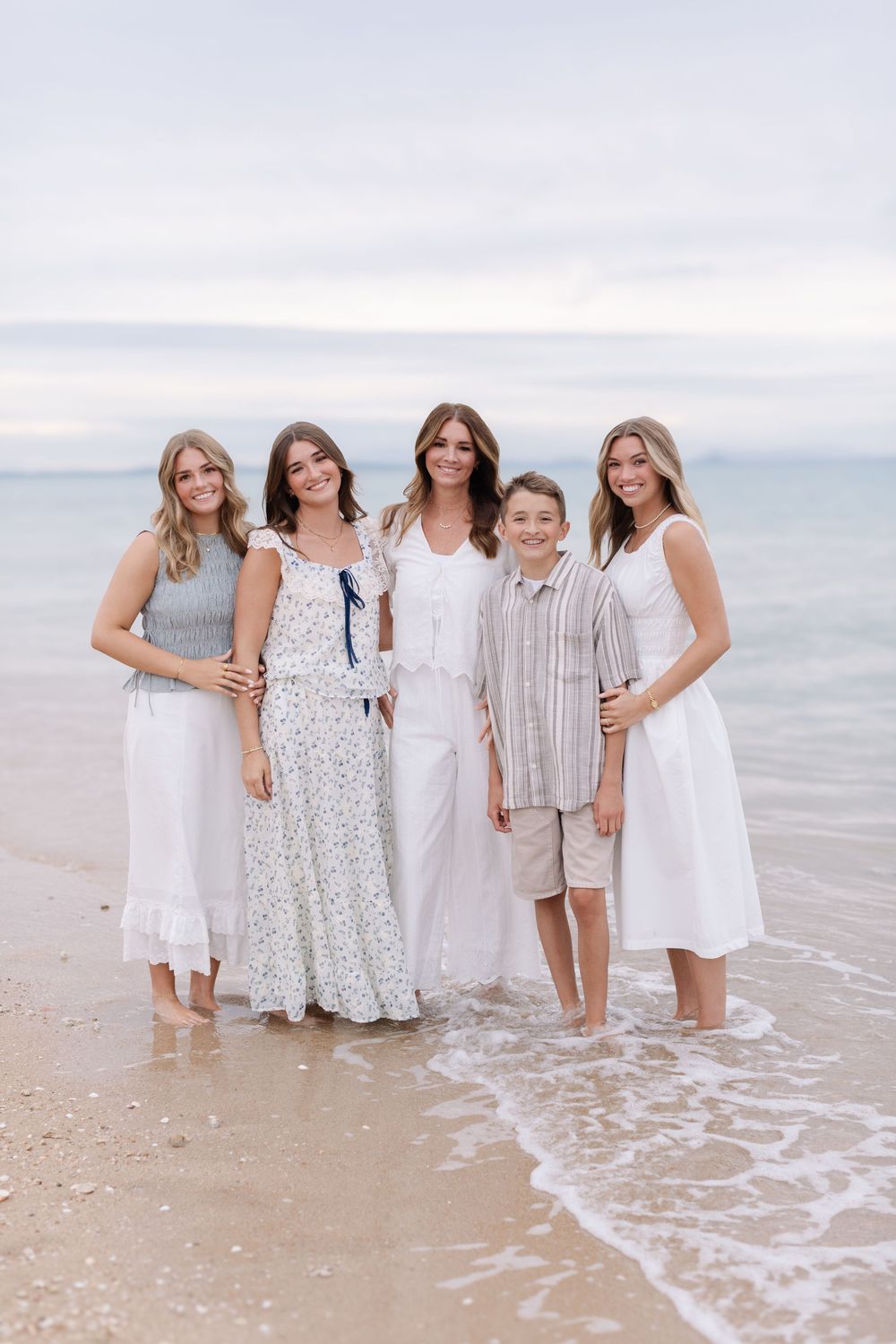 Family of five posing together on a sandy beach shore, wearing coordinated white and neutral outfits, smiling at the camera.