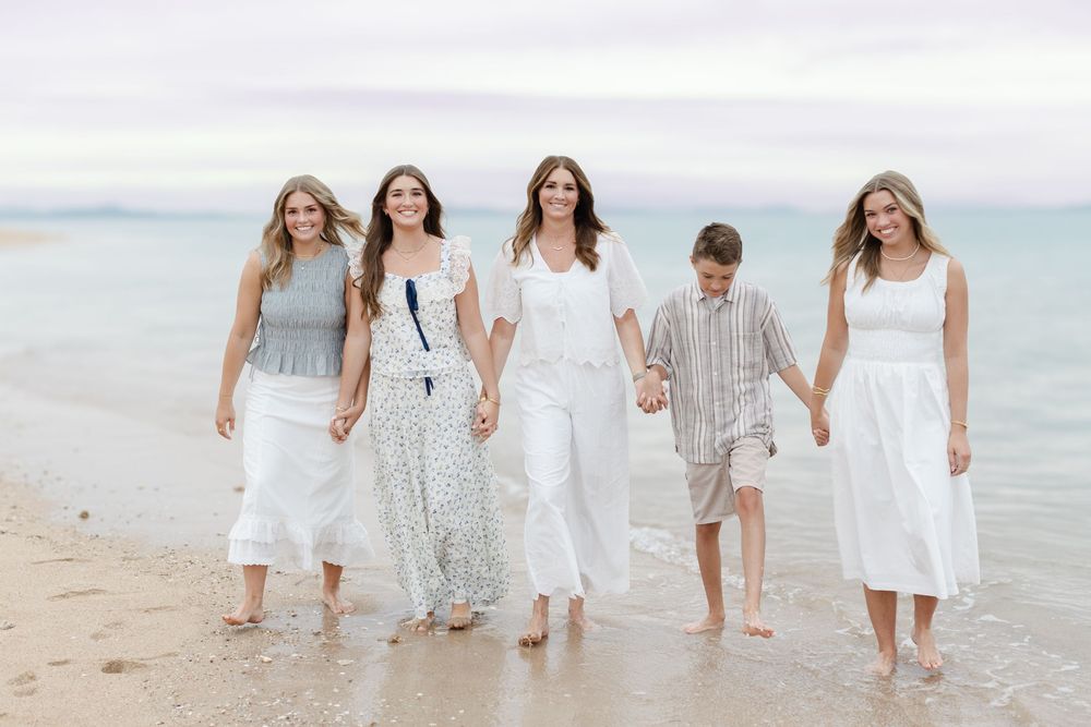 Five people in white outfits walk hand-in-hand along a sandy beach shoreline on a cloudy day.