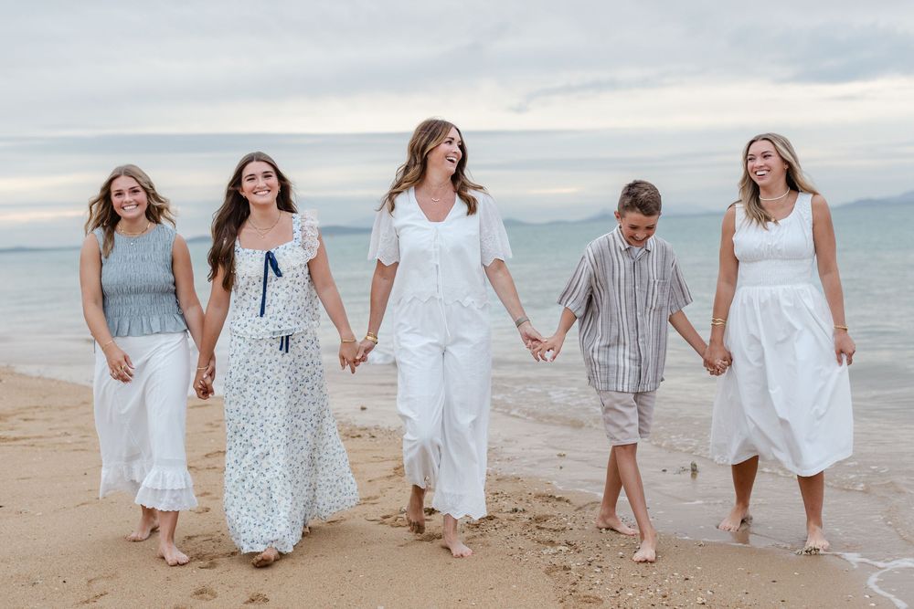 Five people in white outfits walk hand-in-hand on a sandy beach with ocean waves in the background.