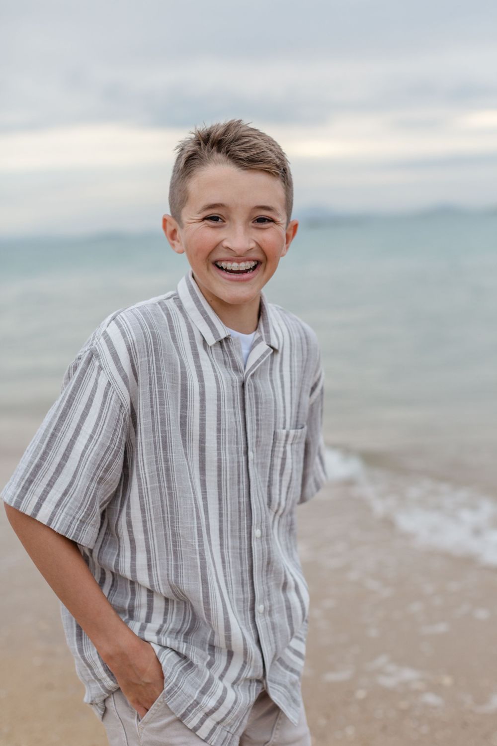 Smiling boy in striped shirt standing on sandy beach with ocean waves in background.