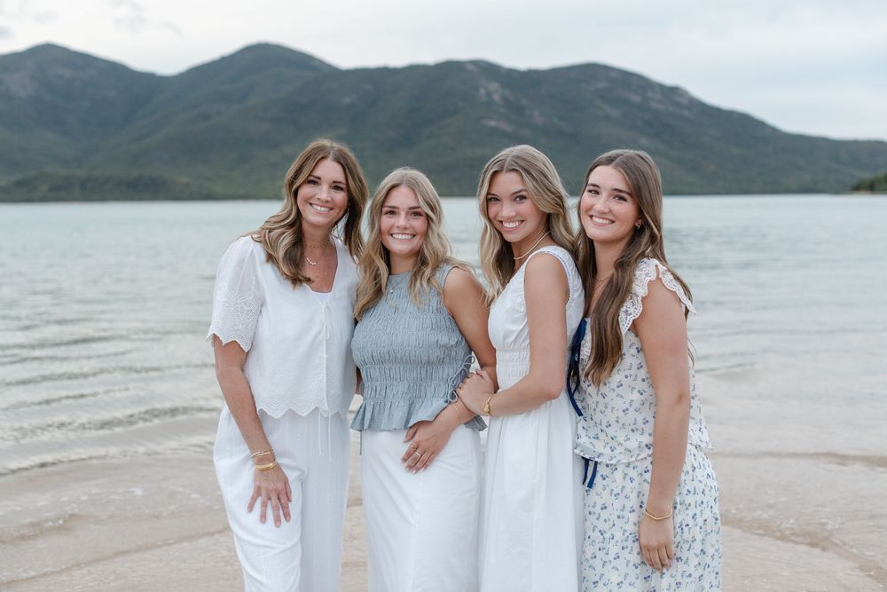 Four women in white and blue outfits smiling together on a lakeside beach with mountains in the background.