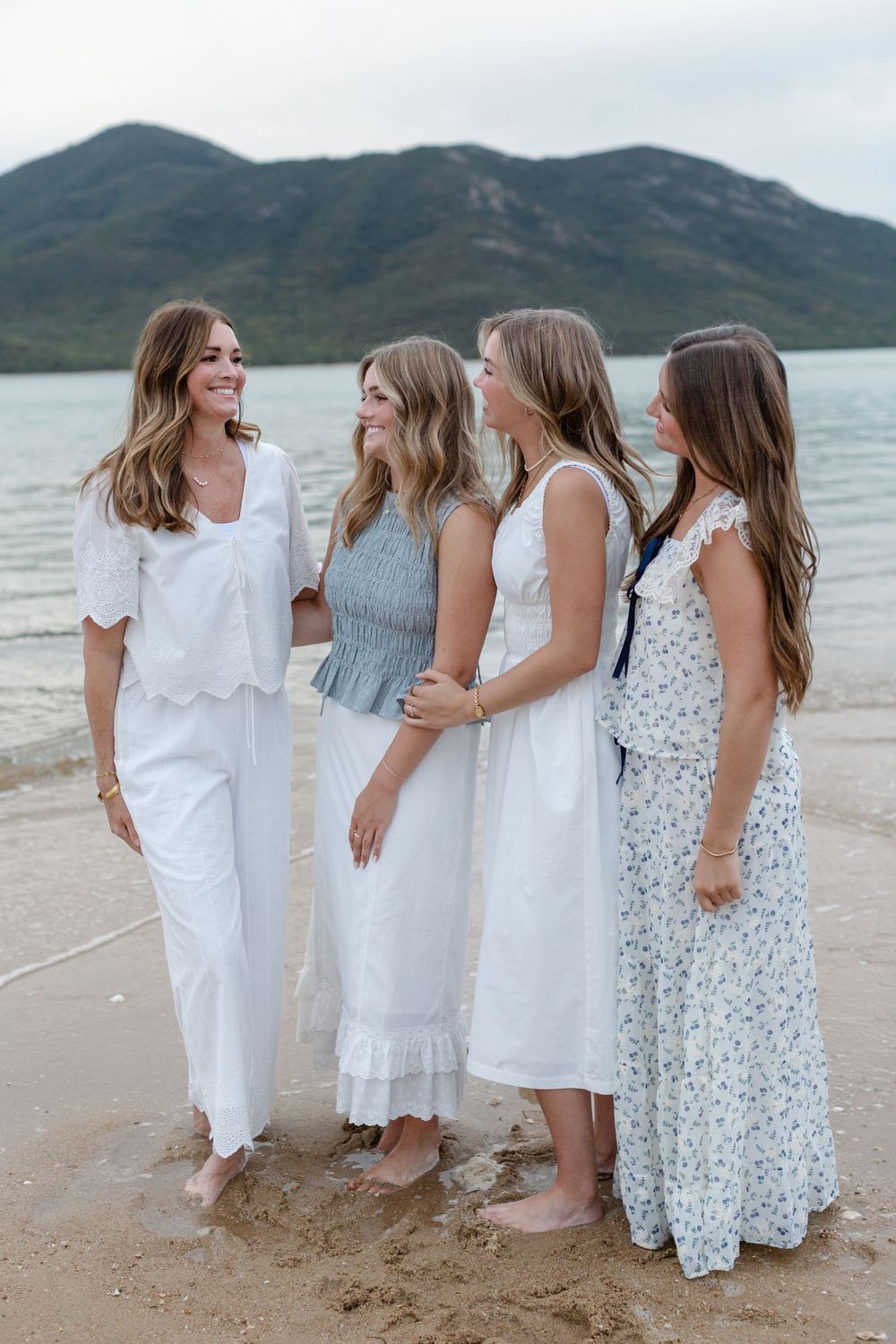 Four women in white and floral dresses standing barefoot on a sandy beach with mountains and water in the background.