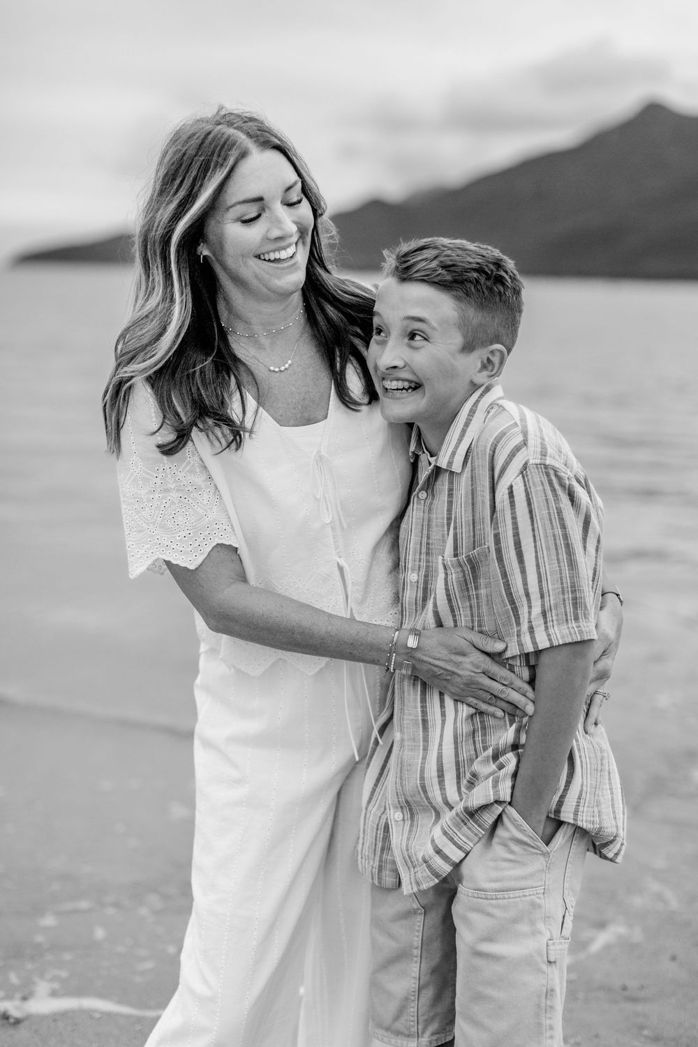 Black and white photo of a smiling mother and son embracing on a beach with mountains in the background.