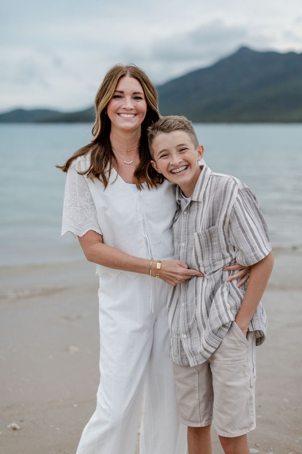 A smiling woman in white and a young boy in striped shirt pose together on a beach with mountains behind.