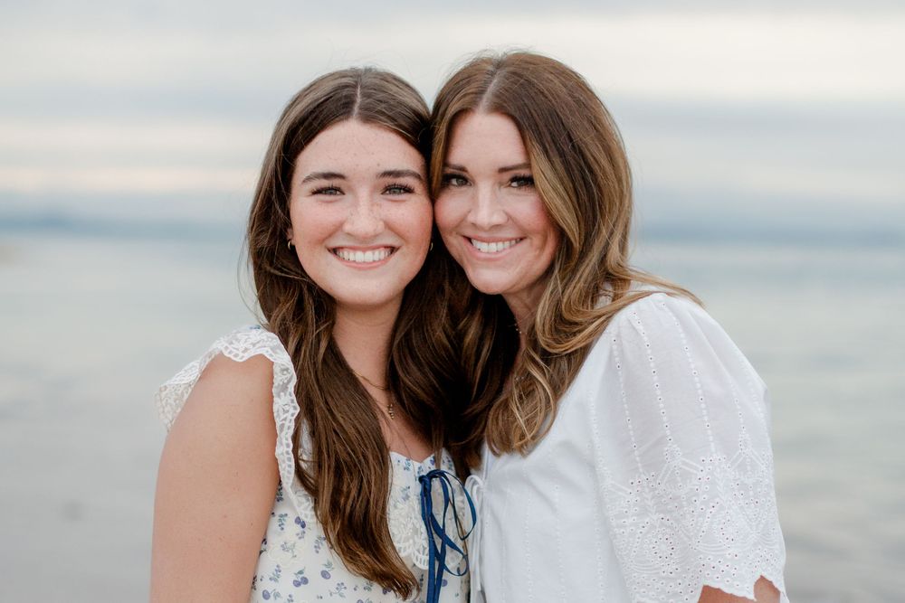 Two smiling young women with brown hair pose together on a beach in white outfits against a soft sky.