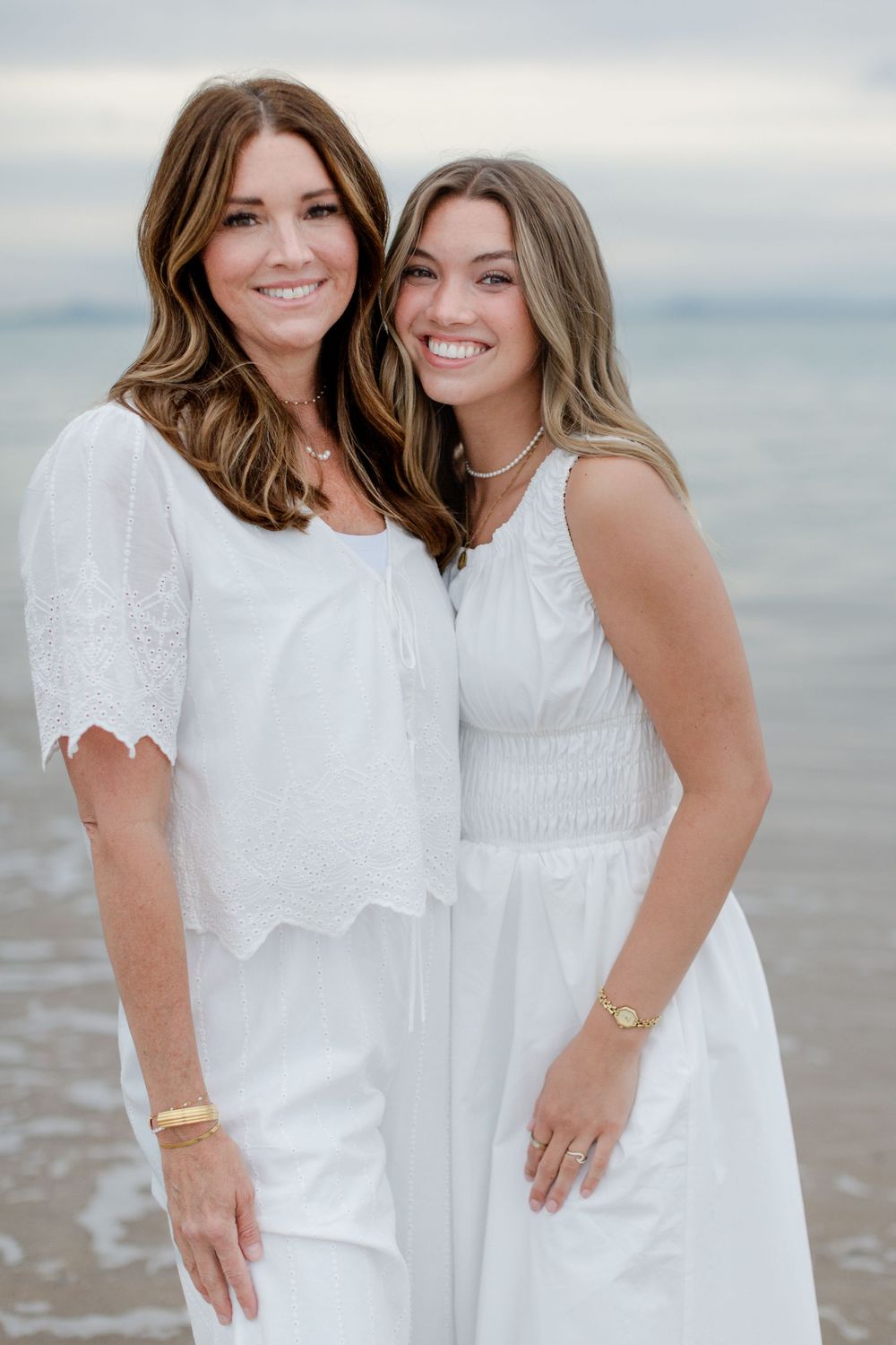 Two women in matching white outfits smiling together on a beach with calm ocean waters in the background.