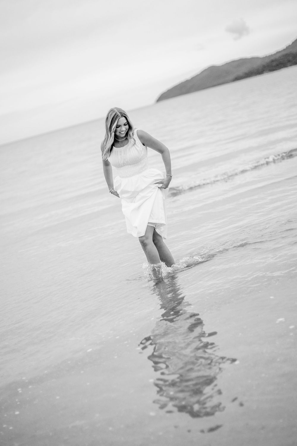 Black and white photo of a woman in a white dress wading in ocean waves on a beach with mountains in the background.