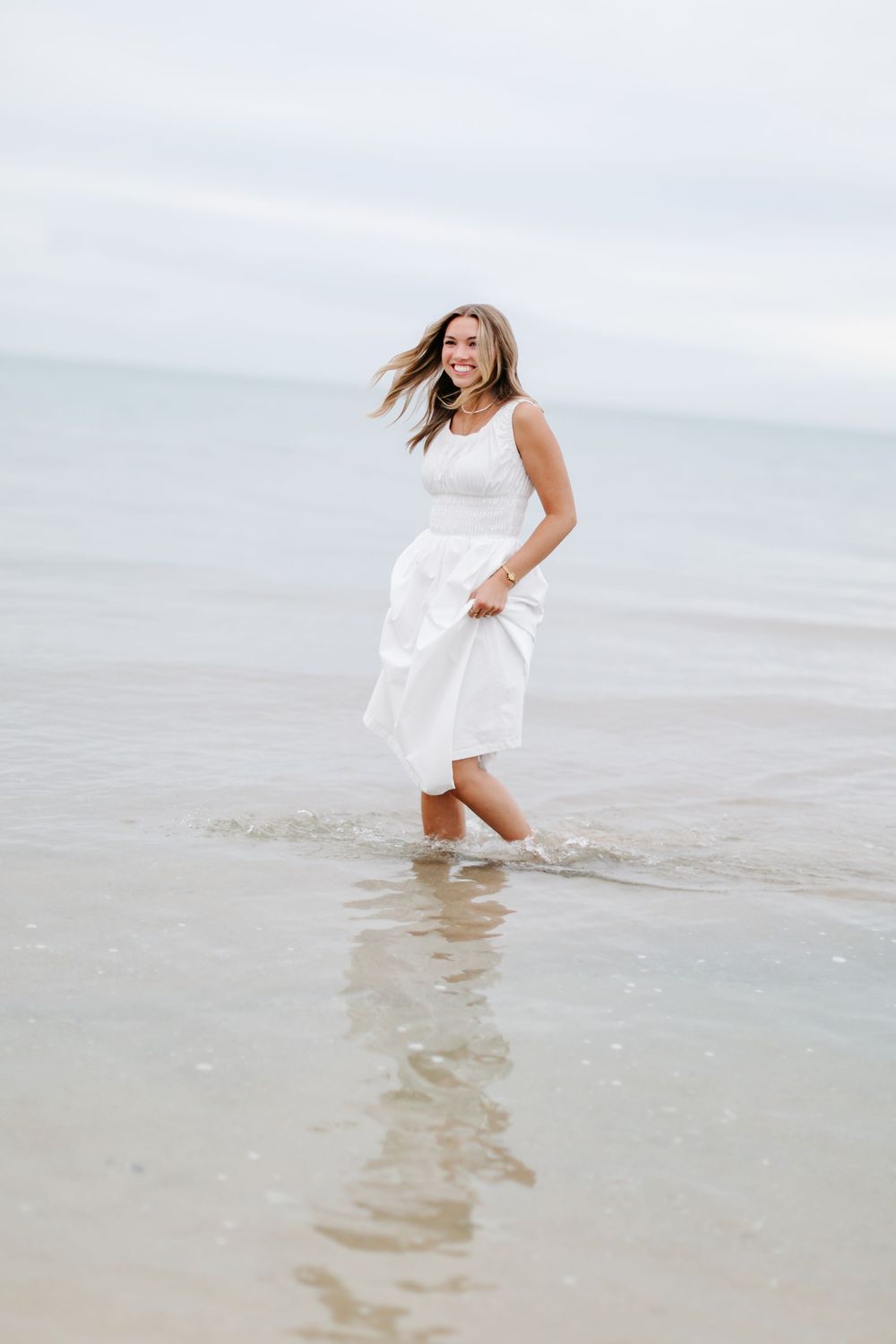A smiling woman in a white dress wading through shallow ocean waves on a beach.