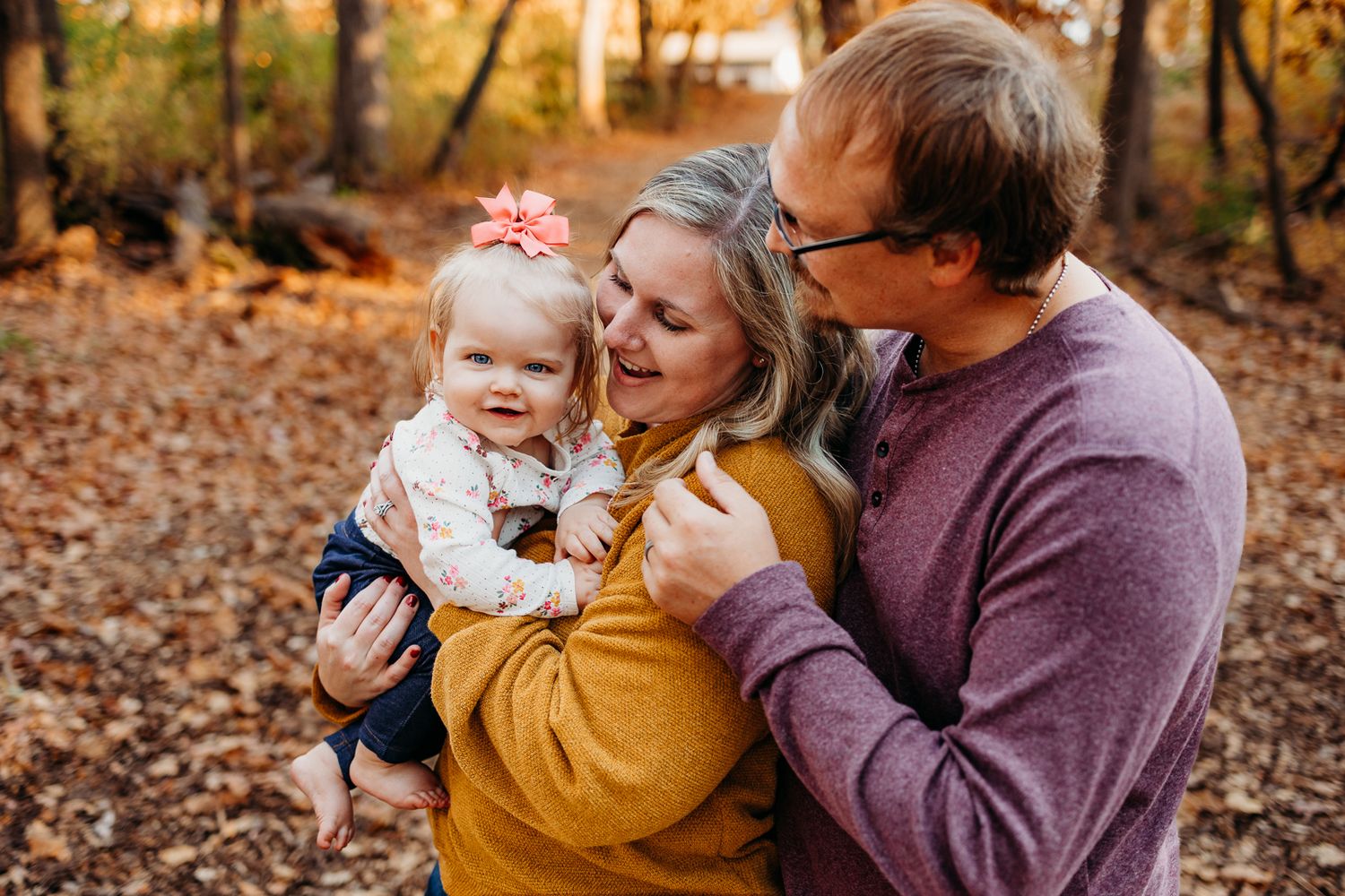 A mom holding a baby girl and the dad resting his hand on the mom's shoulder looking at the baby and smiling. There are leaves on the ground