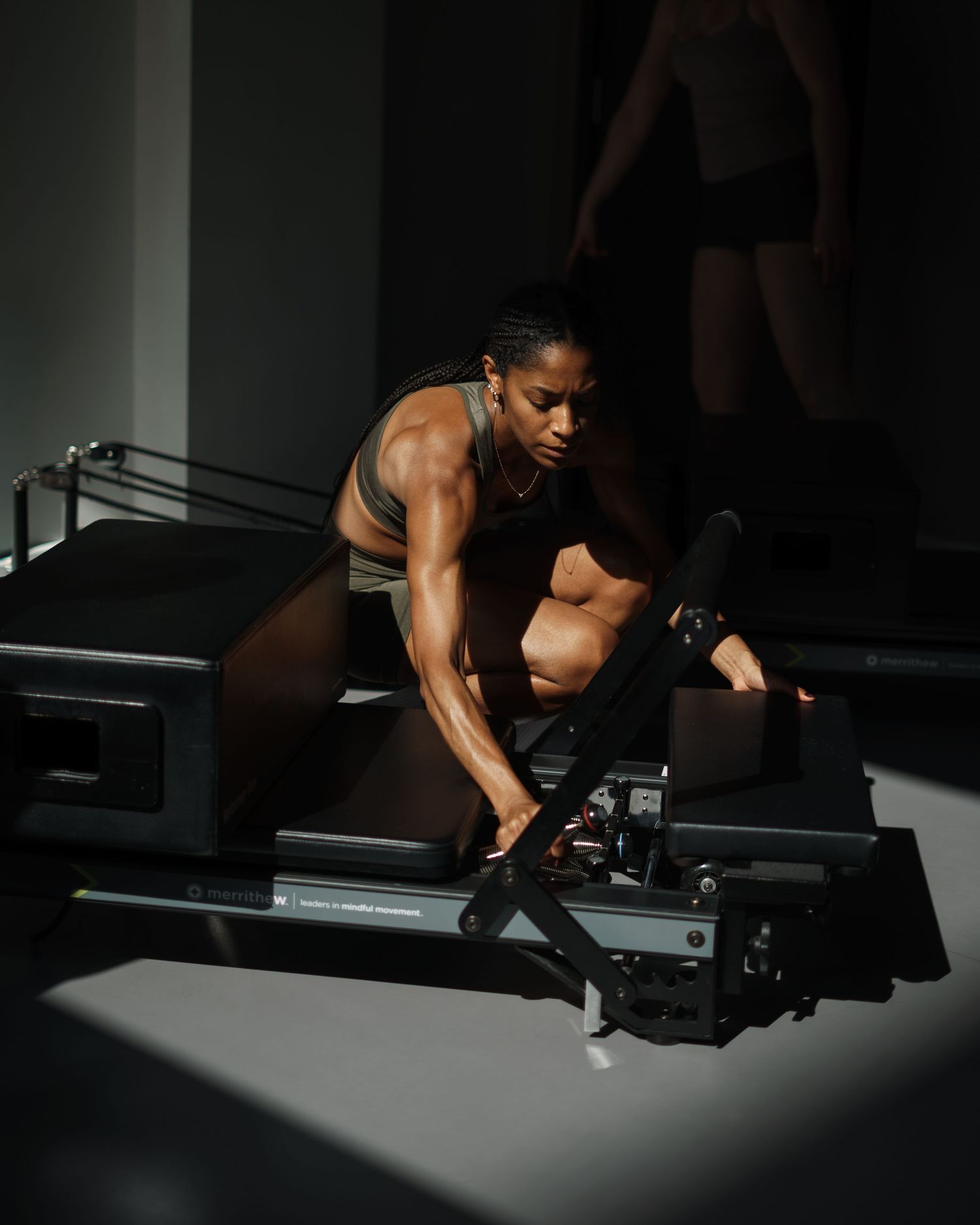 Muscular woman exercises on a Pilates reformer machine in a dramatic, shadow-lit studio setting.