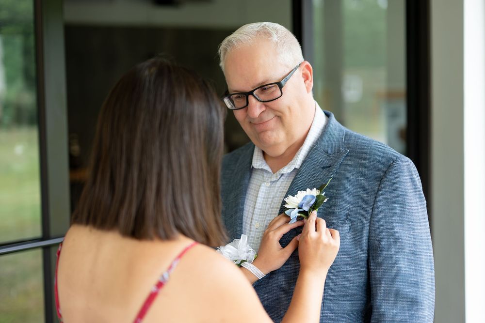 Woman pinning a white boutonniere on a man in a gray blazer and glasses before a wedding ceremony.