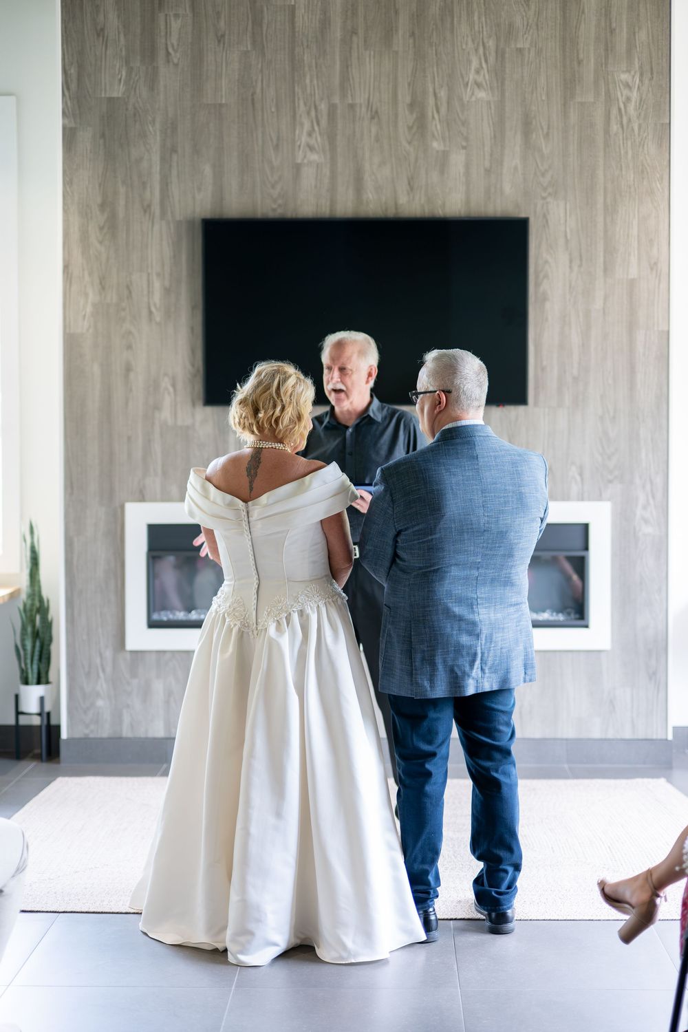 Officiant speaks with bride in white gown and groom in blue suit before fireplace.