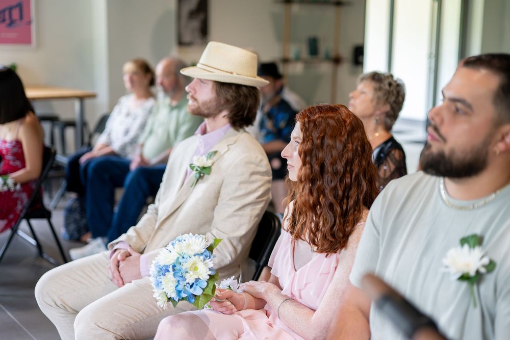 Wedding guests seated at a ceremony, including a man in a straw hat holding a blue floral bouquet.