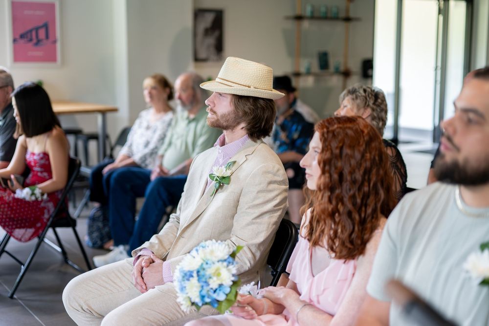A young man in a straw hat holds a blue bouquet beside a redheaded woman in pink at a wedding.