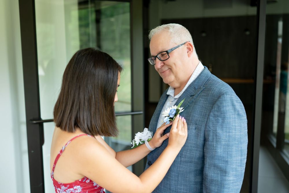 Woman in red floral dress pins boutonniere on groom in blue suit before wedding ceremony.