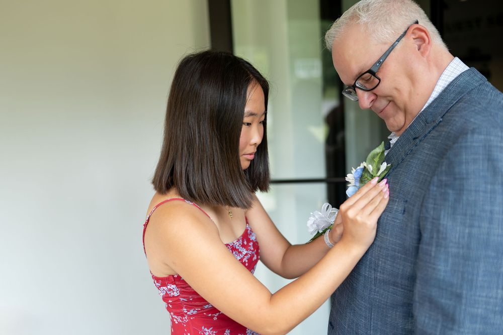 Young girl in red floral dress pins boutonniere on suited man's lapel before wedding ceremony.