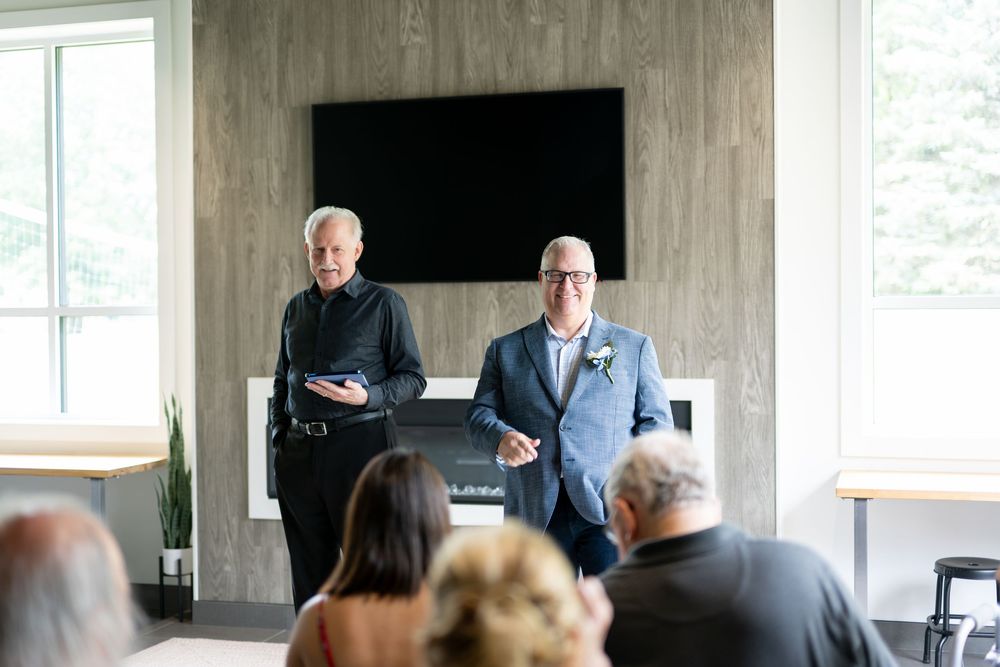 Two men stand before seated wedding guests during an indoor wedding ceremony.