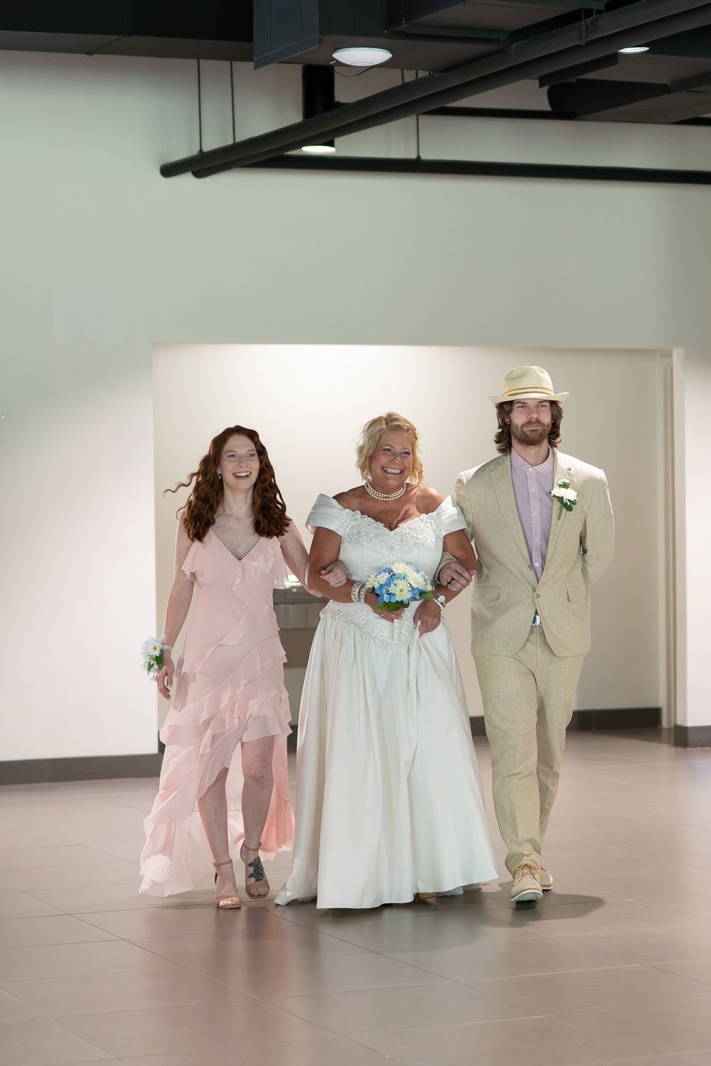 Bride in white gown walks down aisle escorted by redhead woman in pink and man in tan suit and hat.