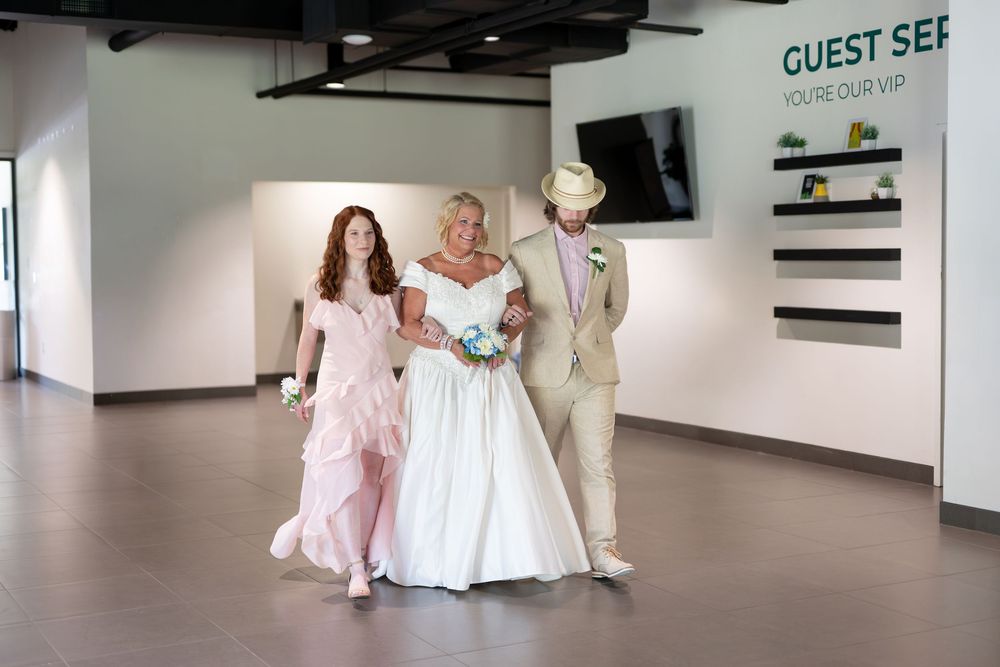 Bride in white gown walks with two guests in pink dress and tan suit through modern venue hallway.