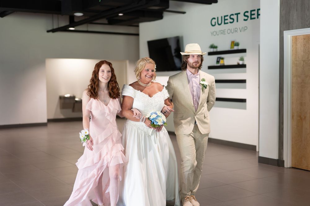 Bride carrying bouquet walks with female and male escorts past guest services desk at wedding venue.