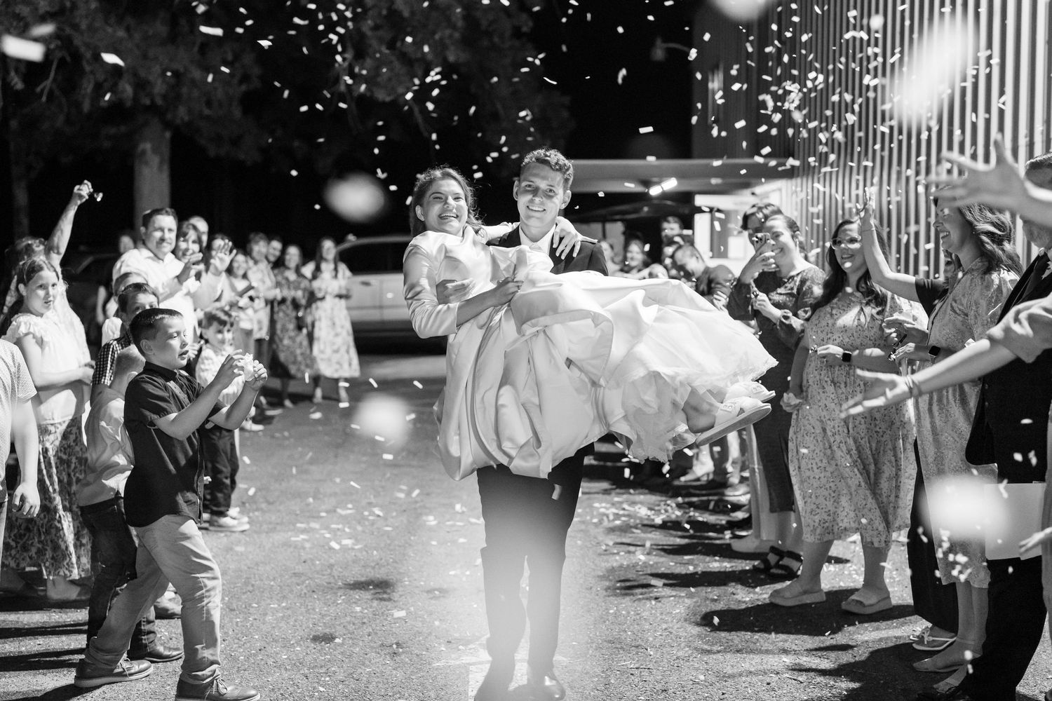 Groom carries bride through confetti shower as guests celebrate wedding send-off in black and white photo.