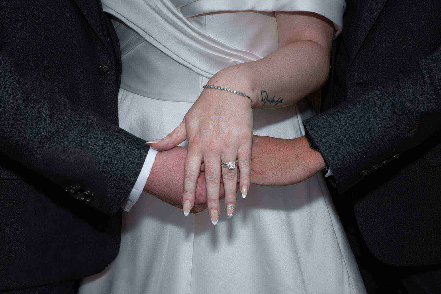 Bride and groom stacking hands showing wedding rings, bride in white dress and groom in dark suit, wedding photography at the den