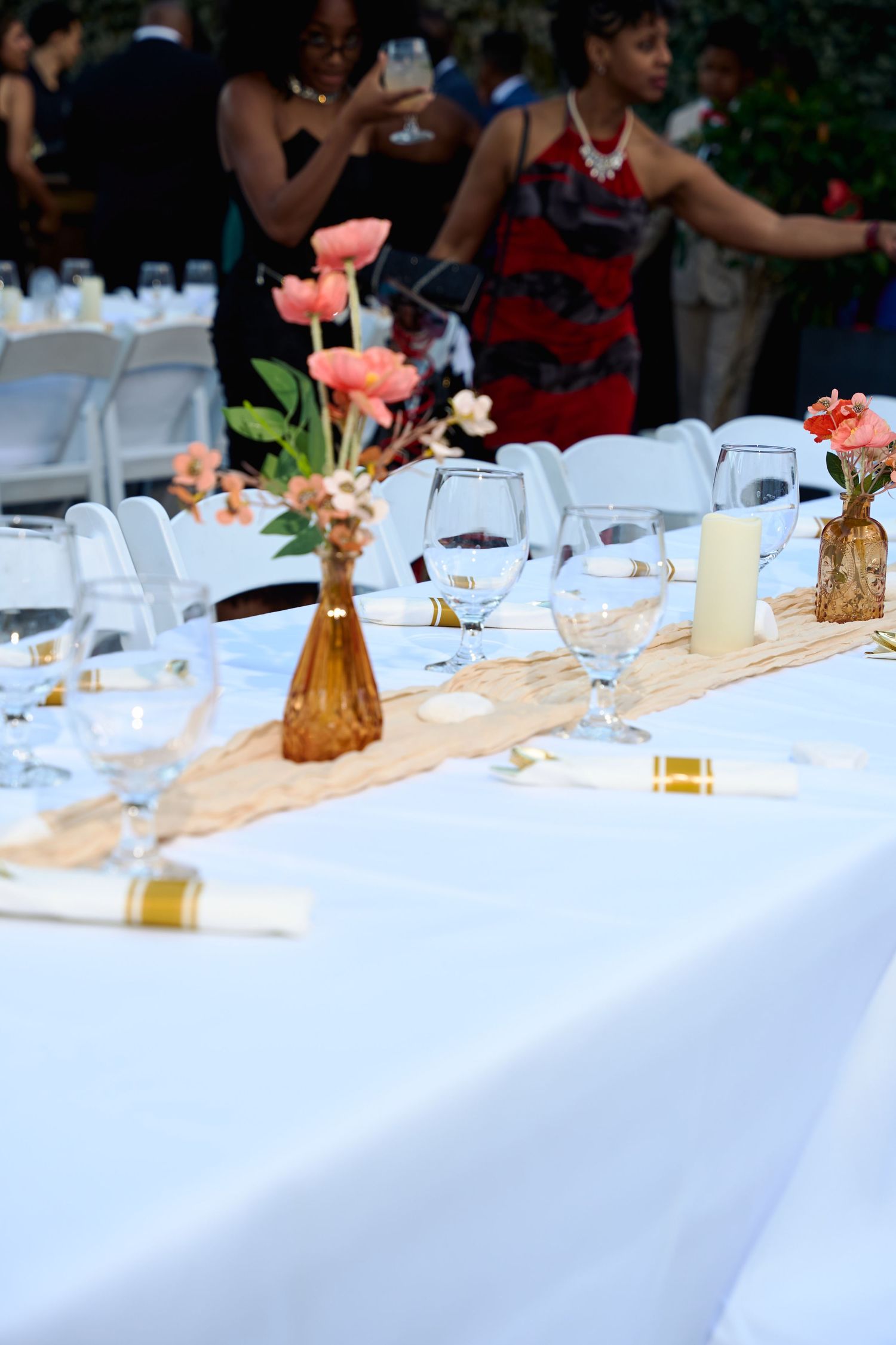 Elegant outdoor wedding reception table set with white linens, coral flowers, gold accents, and crystal glasses.