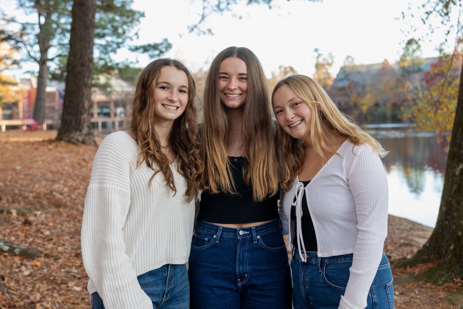 Three smiling young women with long hair pose together outdoors near a lake surrounded by autumn foliage.