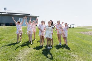 Bridesmaids in matching pink robes jumping joyfully on green grass outside a barn on a sunny day.