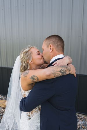Bride and groom sharing a first kiss in wedding attire, bride wearing veil and lace dress with tattoos visible.