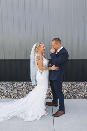 Bride and groom share a tender moment smiling at each other outdoors in wedding attire against a gray barn wall.