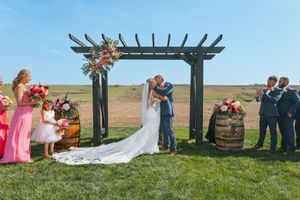 Couple kissing at outdoor wedding ceremony under wooden arbor with floral arrangements, bridesmaids in pink, groomsmen nearby.