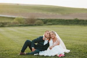 Bride and groom sitting together on lush green grass field, wearing wedding attire, sharing a romantic moment at golden hour.