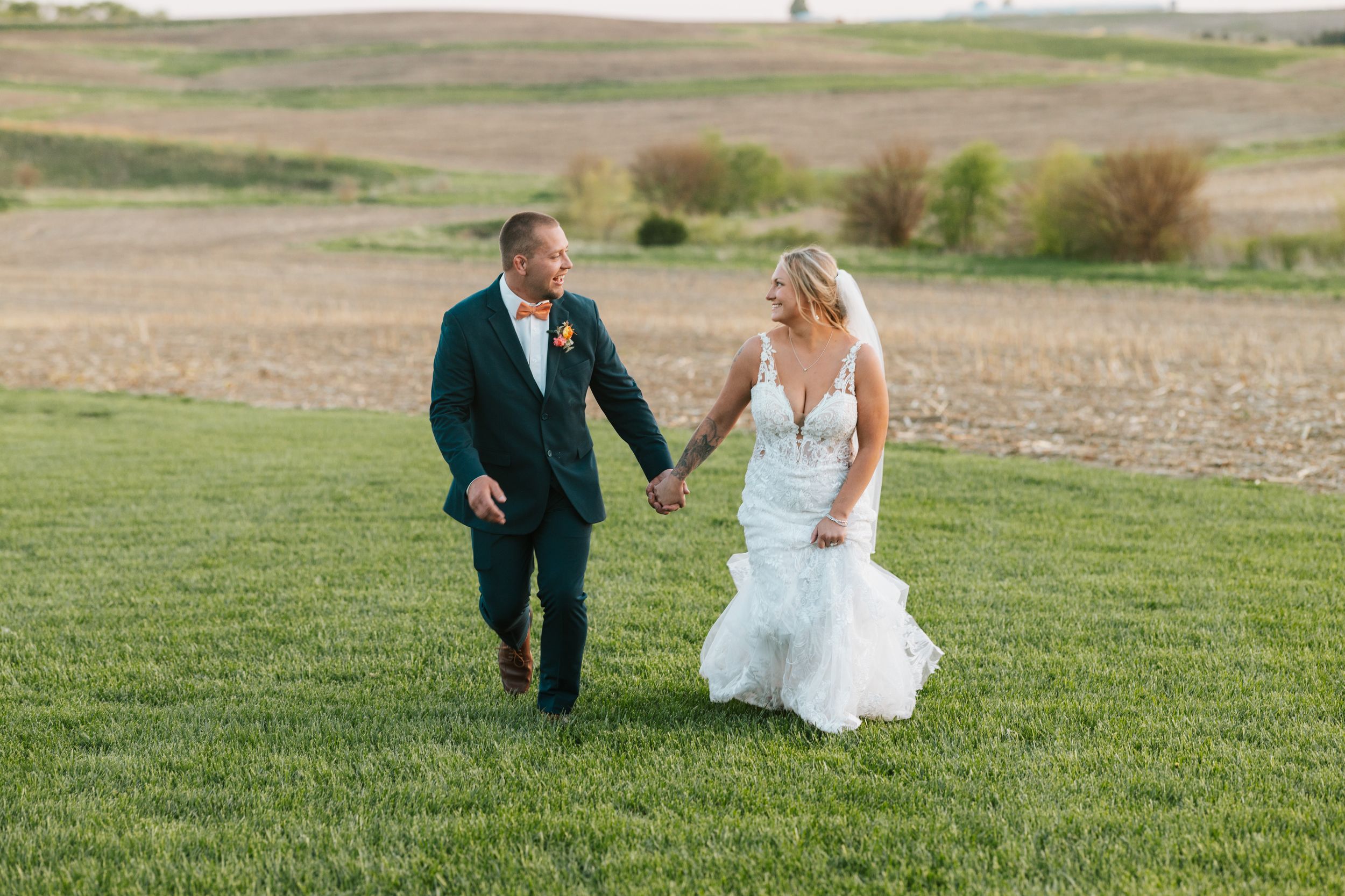Bride and groom holding hands walking through green grass field with rolling farmland hills in background.