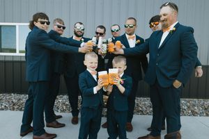 Groomsmen in navy suits toasting with beer glasses outdoors, with two young ring bearers holding drinks at a wedding.