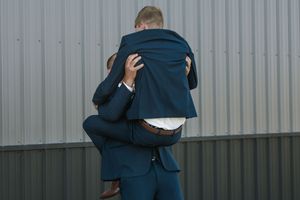 Two men in navy blue suits sharing an embrace outdoors against a corrugated metal wall.