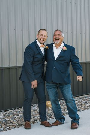 Two men in navy suits laughing together outdoors at a wedding, wearing boutonnieres against a metal barn backdrop.