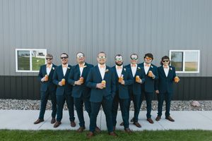 Groomsmen in navy suits and sunglasses holding drinks pose in front of a gray barn wall at a wedding.