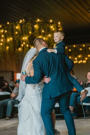 Groom holds young boy in navy suit while dancing with bride under string lights at indoor wedding reception.