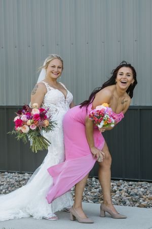 Laughing bride in lace gown playfully poses with bridesmaid in pink dress holding colorful bouquets outside.
