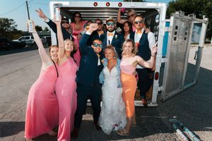 Excited wedding party posing in back of truck, bridesmaids in pink dresses celebrating with raised arms.